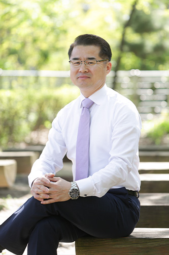 안경과 흰 셔츠, 연보라색 넥타이를 착용한 남성이 야외 벤치에 다리를 꼬고 앉아 있는 모습